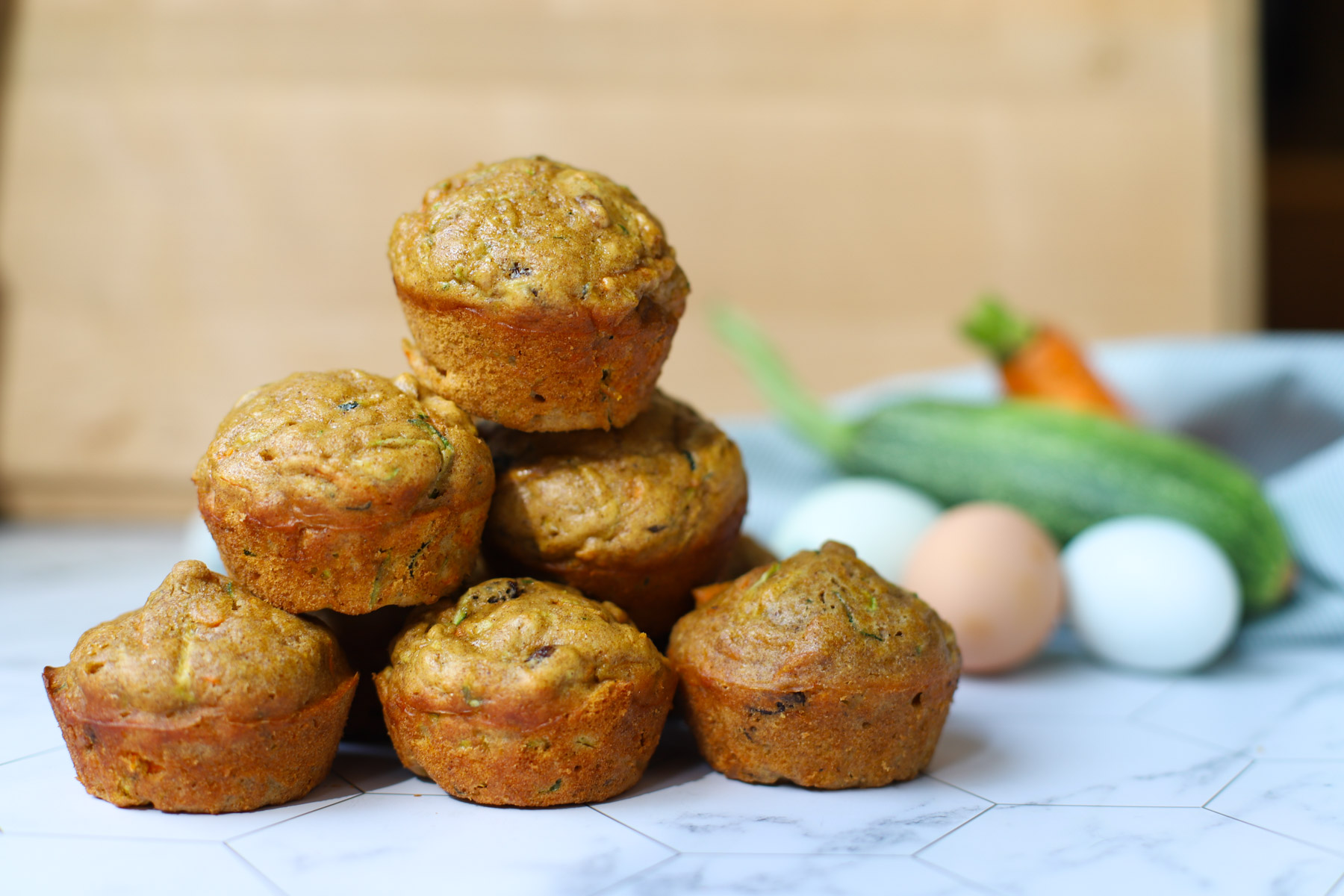 Stack of homemade zucchini carrot muffins on a white marble surface, with fresh zucchini, carrot, and eggs in the background.