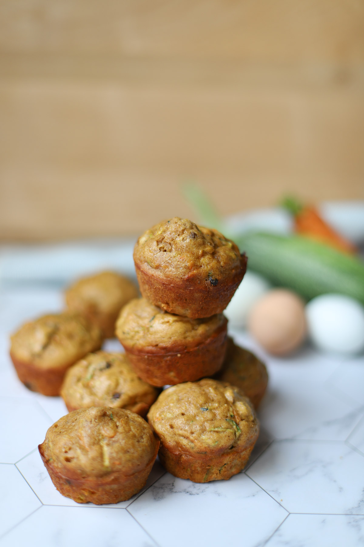 Stack of moist zucchini carrot muffins on a white marble countertop, with fresh vegetables and eggs blurred in the background.