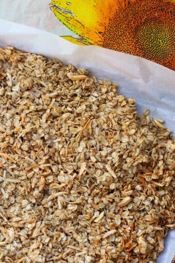 Freshly baked coconut granola with orange zest cooling on a parchment-lined tray, set on a sunflower-patterned cloth.