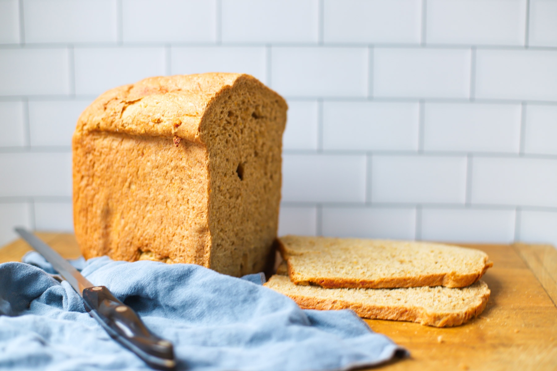 A freshly baked loaf of whole grain rye bread made in a bread machine, with two slices cut and placed on a wooden cutting board, accompanied by a bread knife and a blue linen cloth.