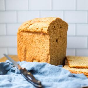 Close-up of a freshly baked whole grain rye bread loaf from a bread machine, resting on a wooden cutting board with a serrated knife and blue linen cloth.