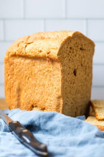Close-up of a freshly baked whole grain rye bread loaf from a bread machine, resting on a wooden cutting board with a serrated knife and blue linen cloth.