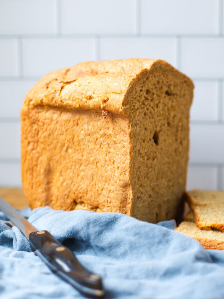 Close-up of a freshly baked whole grain rye bread loaf from a bread machine, resting on a wooden cutting board with a serrated knife and blue linen cloth.