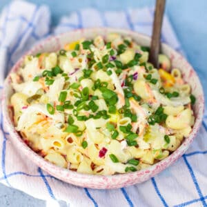 A vibrant bowl of creamy chicken macaroni pasta salad, featuring tender pasta, shredded chicken, crisp coleslaw mix, and a creamy dressing, generously garnished with fresh green chives, set on a blue and white checkered cloth.