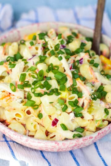 A vibrant bowl of creamy chicken macaroni pasta salad, featuring tender pasta, shredded chicken, crisp coleslaw mix, and a creamy dressing, generously garnished with fresh green chives, set on a blue and white checkered cloth.