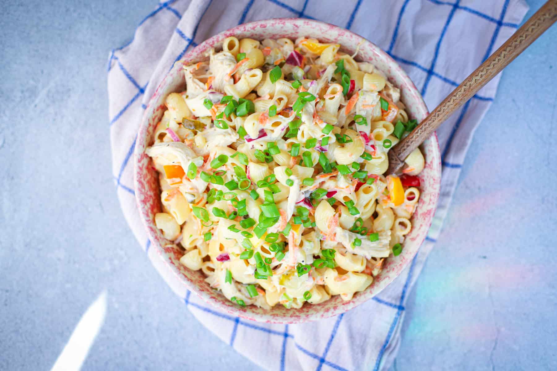 Overhead shot of a colorful chicken macaroni salad with bell peppers, carrots, and green onions in a pink speckled bowl on a blue towel.