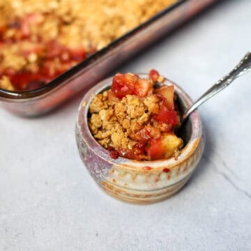 Homemade apple blackberry crumble served in a rustic ceramic bowl with a spoon, with the baked crumble dish in the background.