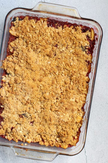 A delicious, golden-brown apple and blackberry crumble in a clear glass baking dish, sitting on a light gray countertop. The topping is a mix of toasted oats and flour, and the bubbling fruit filling is visible along the edges.