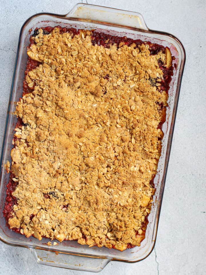 A delicious, golden-brown apple and blackberry crumble in a clear glass baking dish, sitting on a light gray countertop. The topping is a mix of toasted oats and flour, and the bubbling fruit filling is visible along the edges.