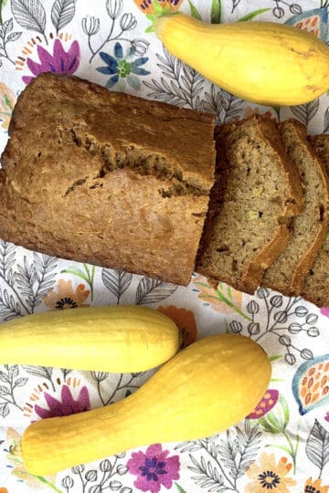Top-down view of a golden-brown, sliced loaf of homemade yellow squash bread displayed with three whole yellow summer squash on a bright, floral tea towel, serving as the featured image for the recipe.
