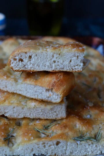 A stack of three sliced squares of thick, airy sourdough discard focaccia bread showing a porous crumb and golden crust topped with fresh rosemary and coarse sea salt.