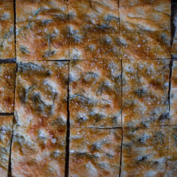 An overhead shot of a tray of golden-brown sourdough discard focaccia sliced into squares, topped with coarse sea salt and fresh rosemary needles.