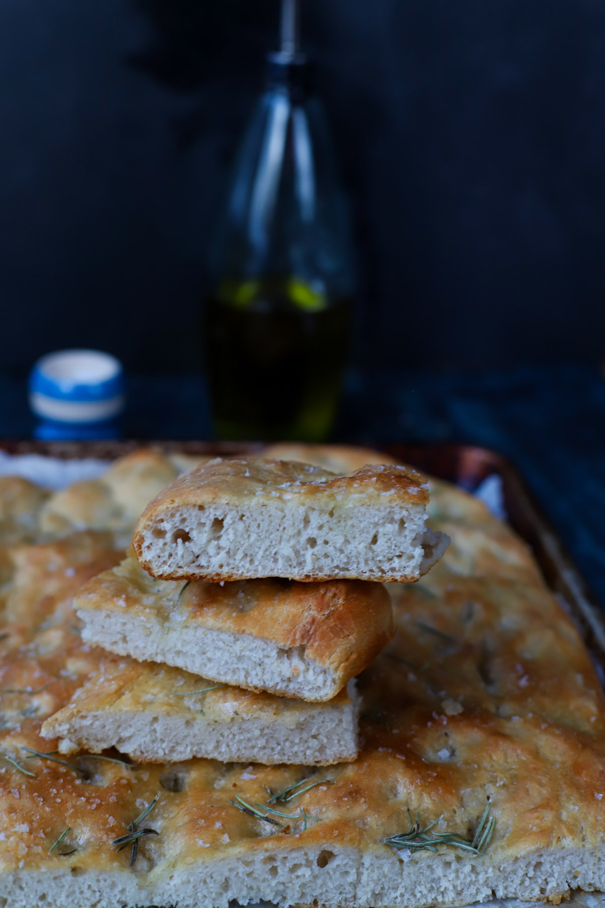 Three slices of homemade sourdough discard focaccia stacked to show the airy crumb, with a glass olive oil bottle and salt cellar in the blurred background.