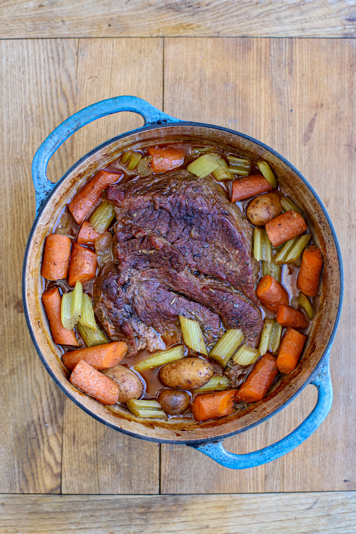 Top-down view of a succulent braised chuck roast swimming in rich gravy with carrots and celery inside a rustic blue Dutch oven on a wooden table.
