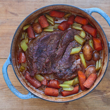 An overhead view of a hearty beef pot roast with thick-cut carrots, celery, and baby potatoes in a savory broth inside a blue enameled Dutch oven.