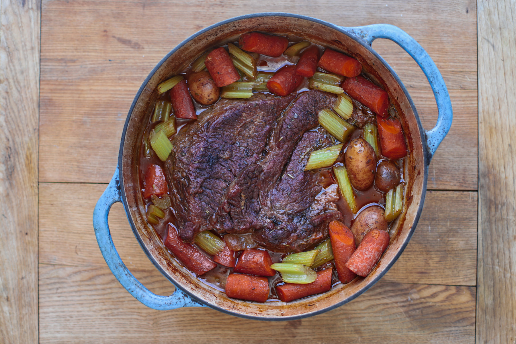 An overhead view of a hearty beef pot roast with thick-cut carrots, celery, and baby potatoes in a savory broth inside a blue enameled Dutch oven.