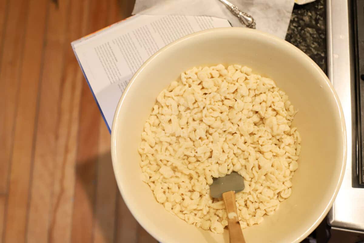 Cooked spaetzle in a cream mixing bowl with a silicone spatula, with an open cookbook on the counter beside it