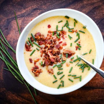 Bowl of loaded potato soup with bacon and chives beside a bundle of fresh chives on a rustic wood surface