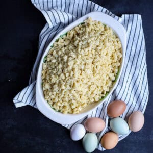 Overhead view of homemade spaetzle in a white oval baking dish next to colorful fresh eggs on a striped towel