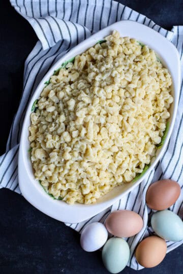 Overhead view of homemade spaetzle in a white oval baking dish next to colorful fresh eggs on a striped towel